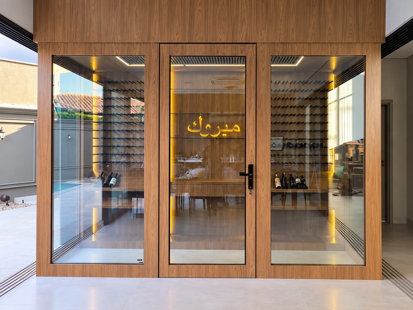 Sala de vinhos com fachada de vidro emoldurada em madeira, porta central, iluminação em LED e suportes de garrafa nas paredes internas