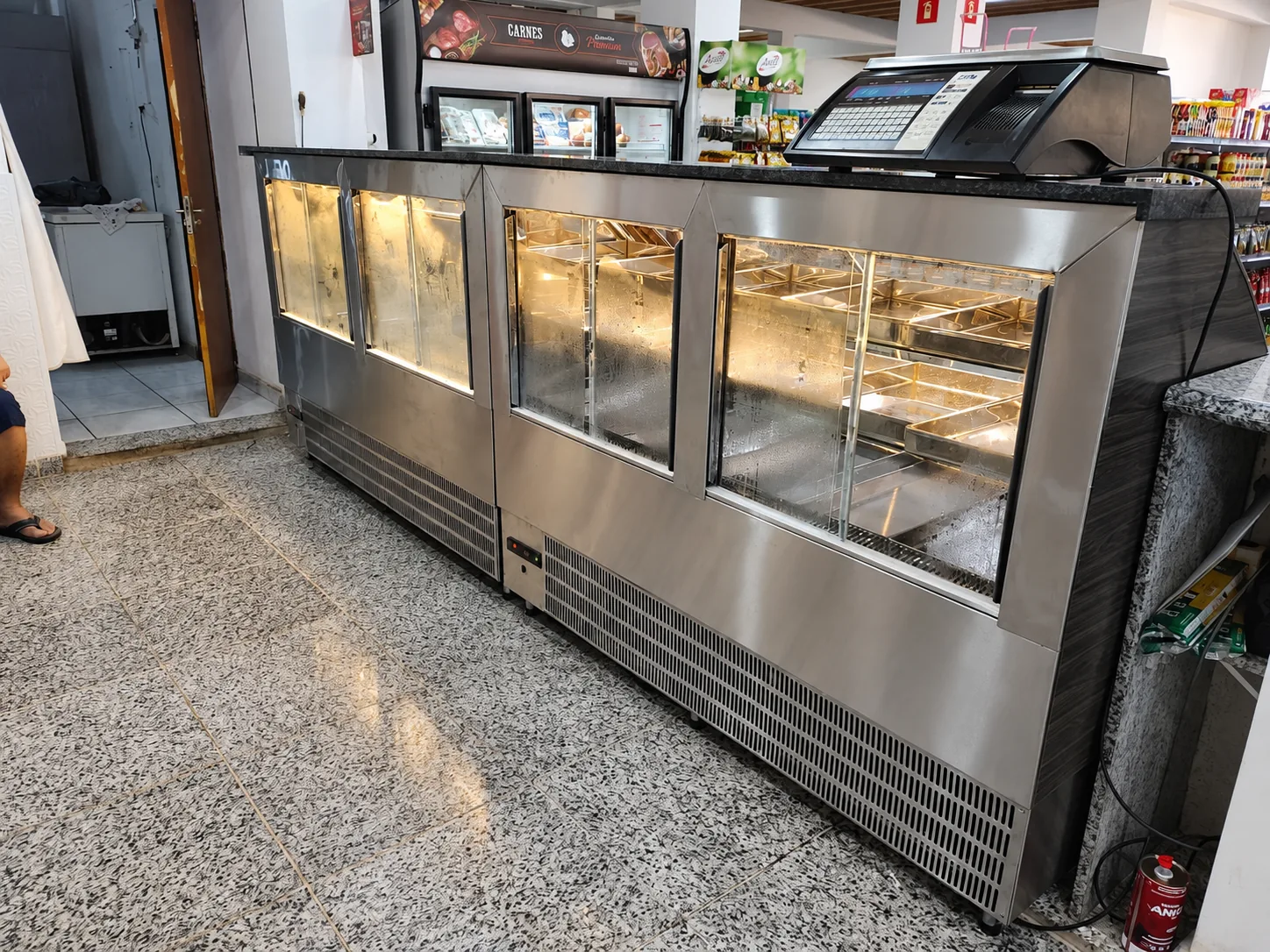 Balcão refrigerado em inox escovado com portas de vidro iluminadas, grelhas de ventilação na base e balança sobre o tampo de granito