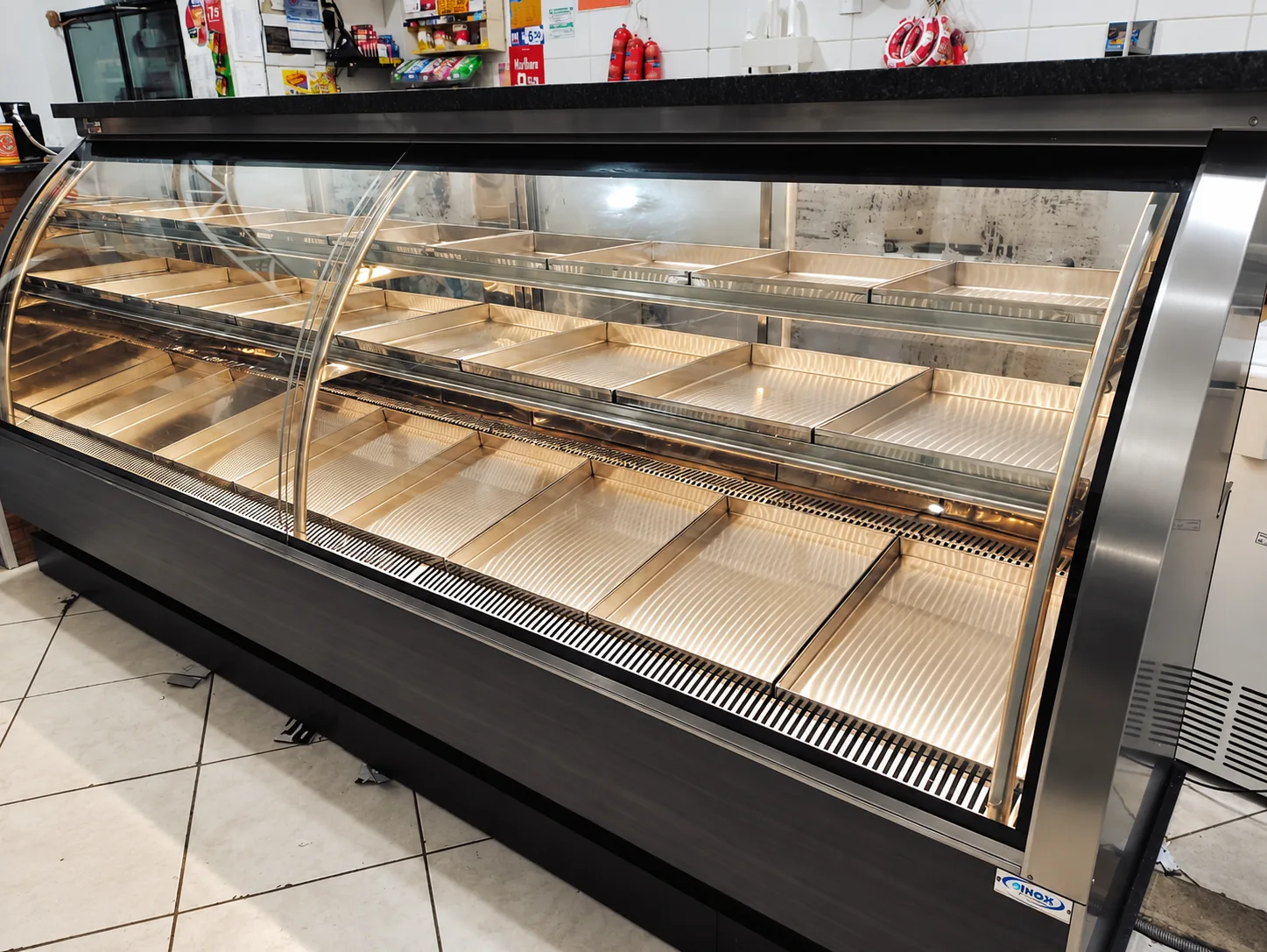 Balcão expositor refrigerado vazio, com vidro curvo, iluminação interna e bandejas em inox organizadas em três níveis, frente em madeira escura
