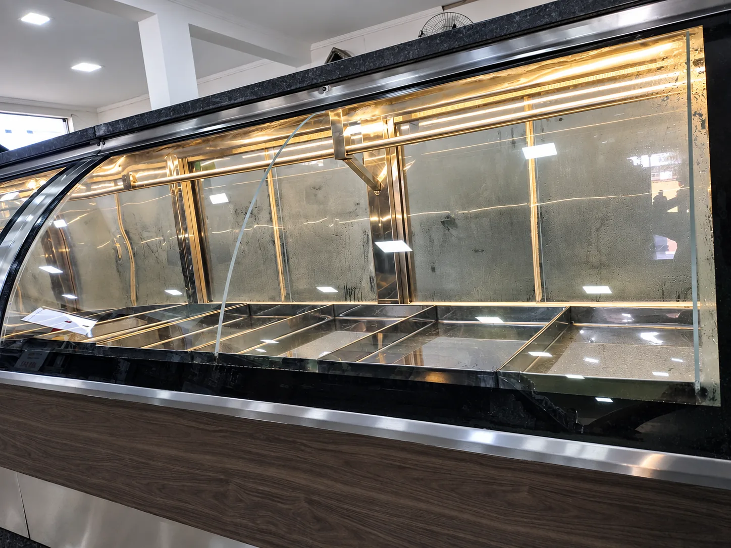 Detalhe de balcão expositor de açougue com vidro curvo, perfil em inox, iluminação interna em LED e bandejas em inox, frente em madeira