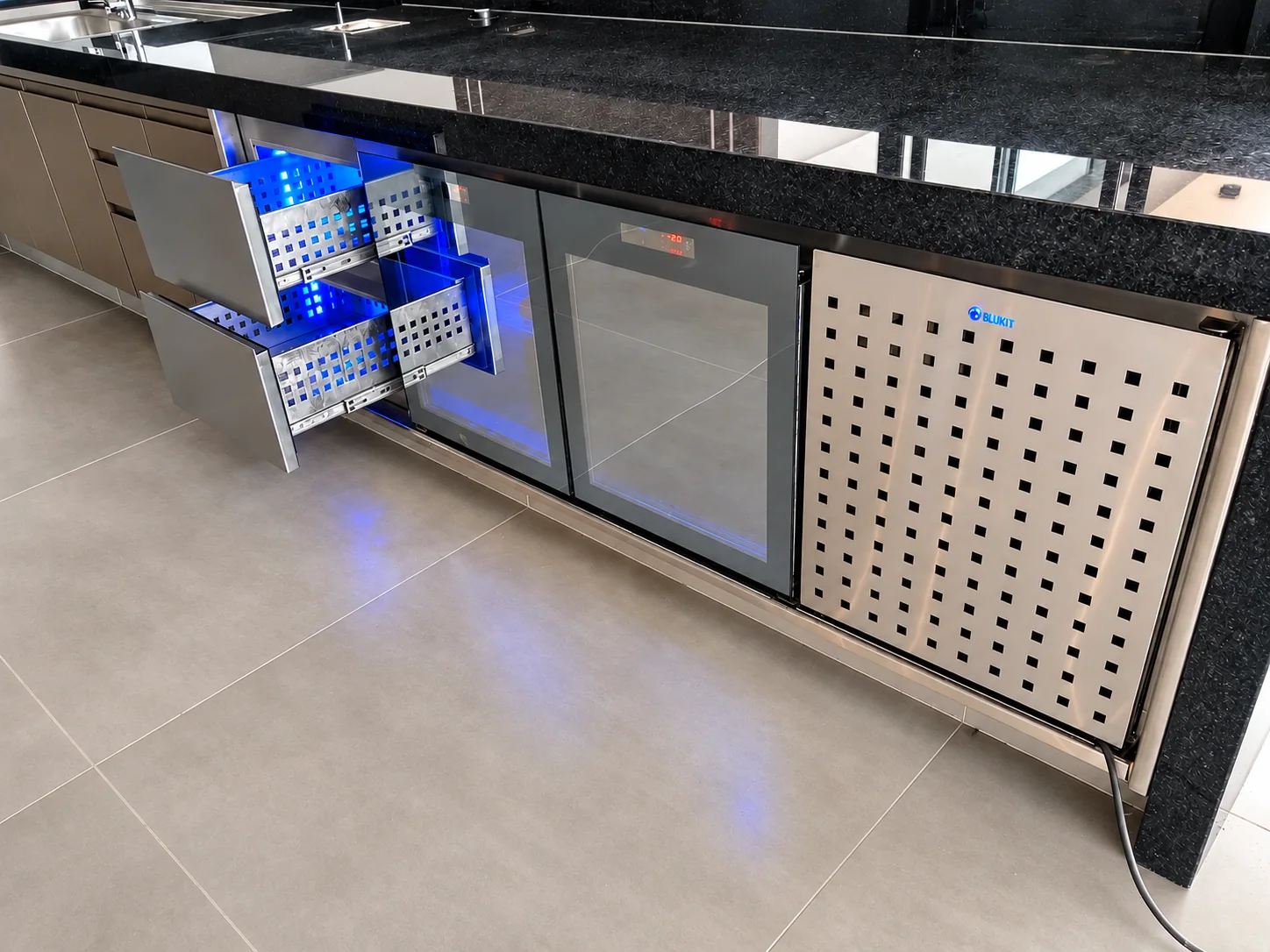 Balcão gourmet com tampo de granito preto, gavetas refrigeradas com cestos em inox e luz azul, duas portas de vidro e painel perfurado em inox de ventilação