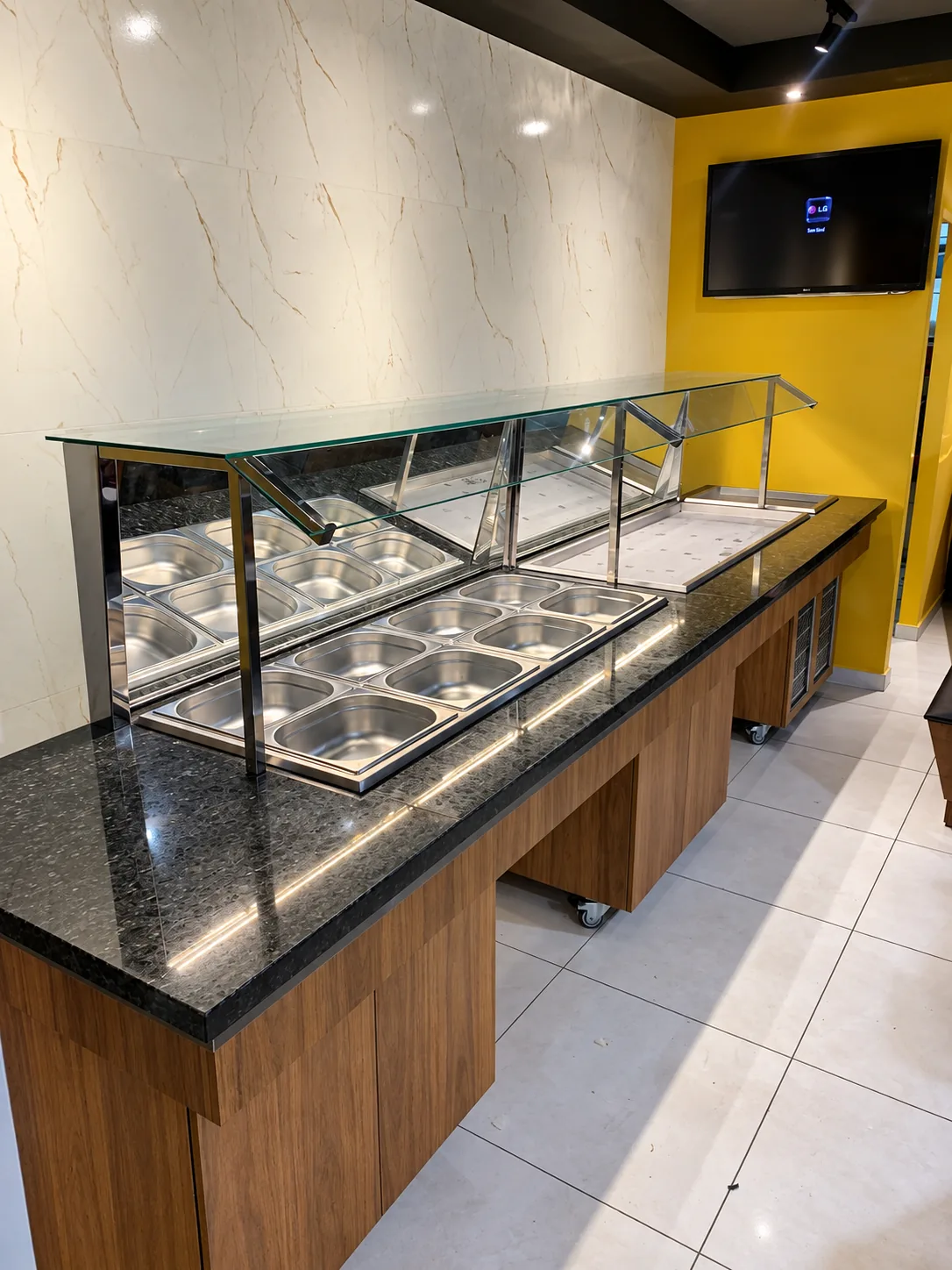 Buffet com cubas GN em inox embutidas no tampo de granito, protetor de vidro com estrutura em inox e base em madeira sobre rodízios