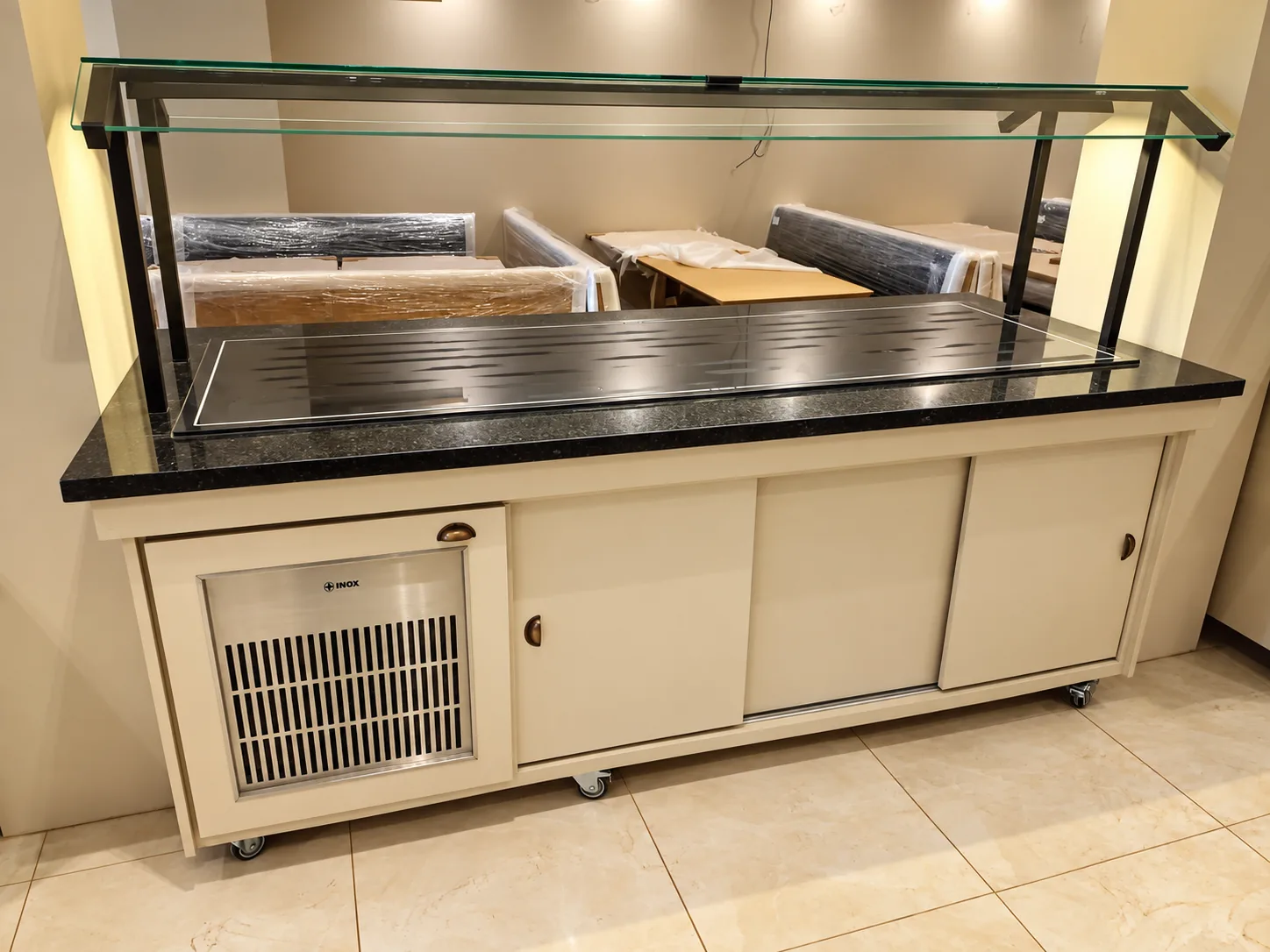 Buffet refrigerado com tampo de granito, protetor de vidro com estrutura preta, portas de correr na cor do ambiente e unidade de refrigeração com a marca Mais Inox, sobre rodízios