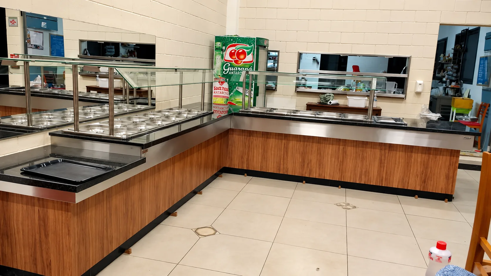 Linha de buffet em L com cubas GN em inox sob protetor de vidro, tampo de granito, faixa em inox e frente em madeira, com apoio de bandejas na entrada