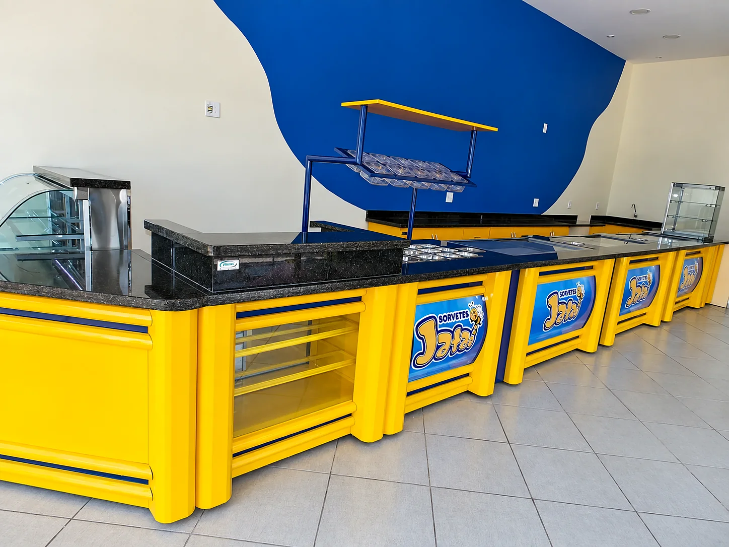 Balcão de sorveteria com vitrine curva, prateleira aérea de casquinhas e cubas refrigeradas