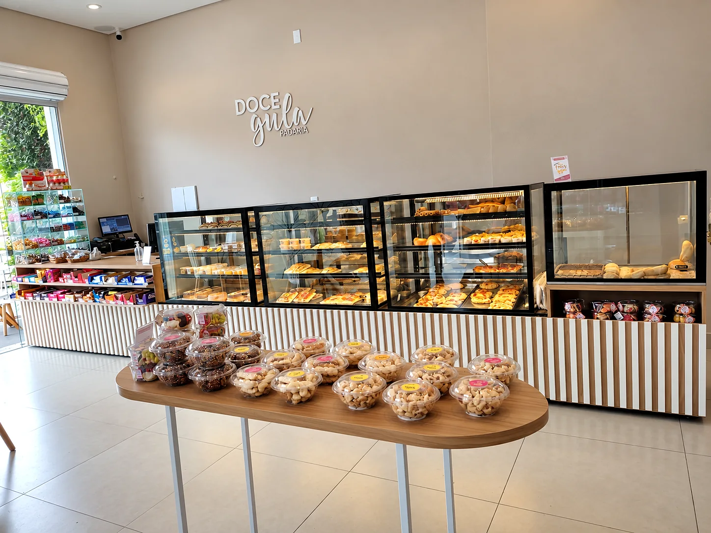 Salão de padaria com linha de vitrines refrigeradas iluminadas, balcão de caixa e mesa de apoio com potes de biscoitos