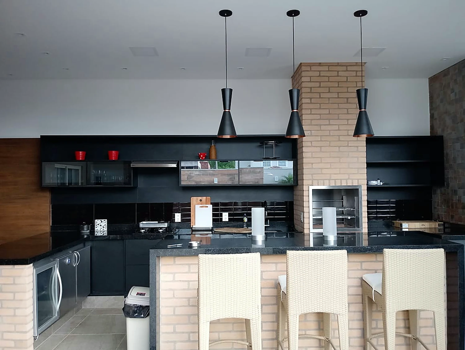 Área gourmet com churrasqueira embutida em alvenaria de tijolo, bancada de granito, banquetas e armários escuros