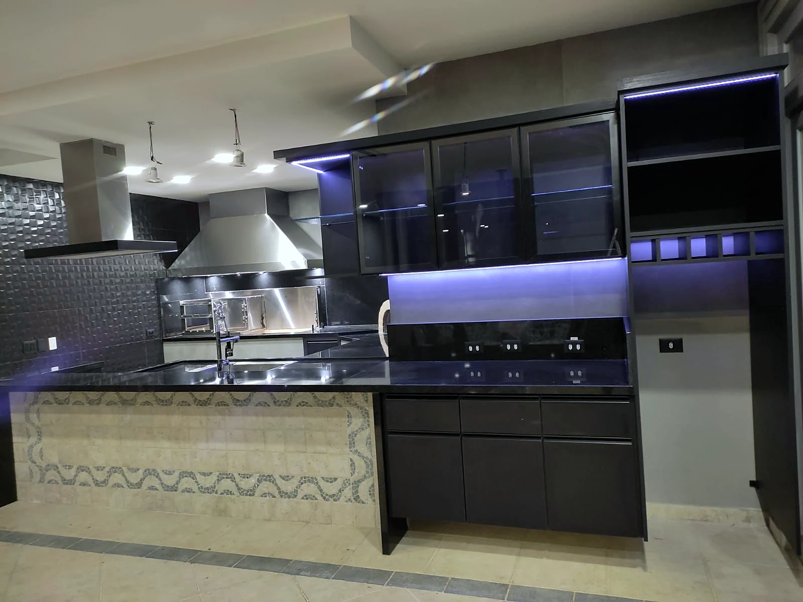 Área gourmet com coifa e churrasqueira em inox, armários pretos com portas de vidro e iluminação embutida, e balcão com frente em mosaico
