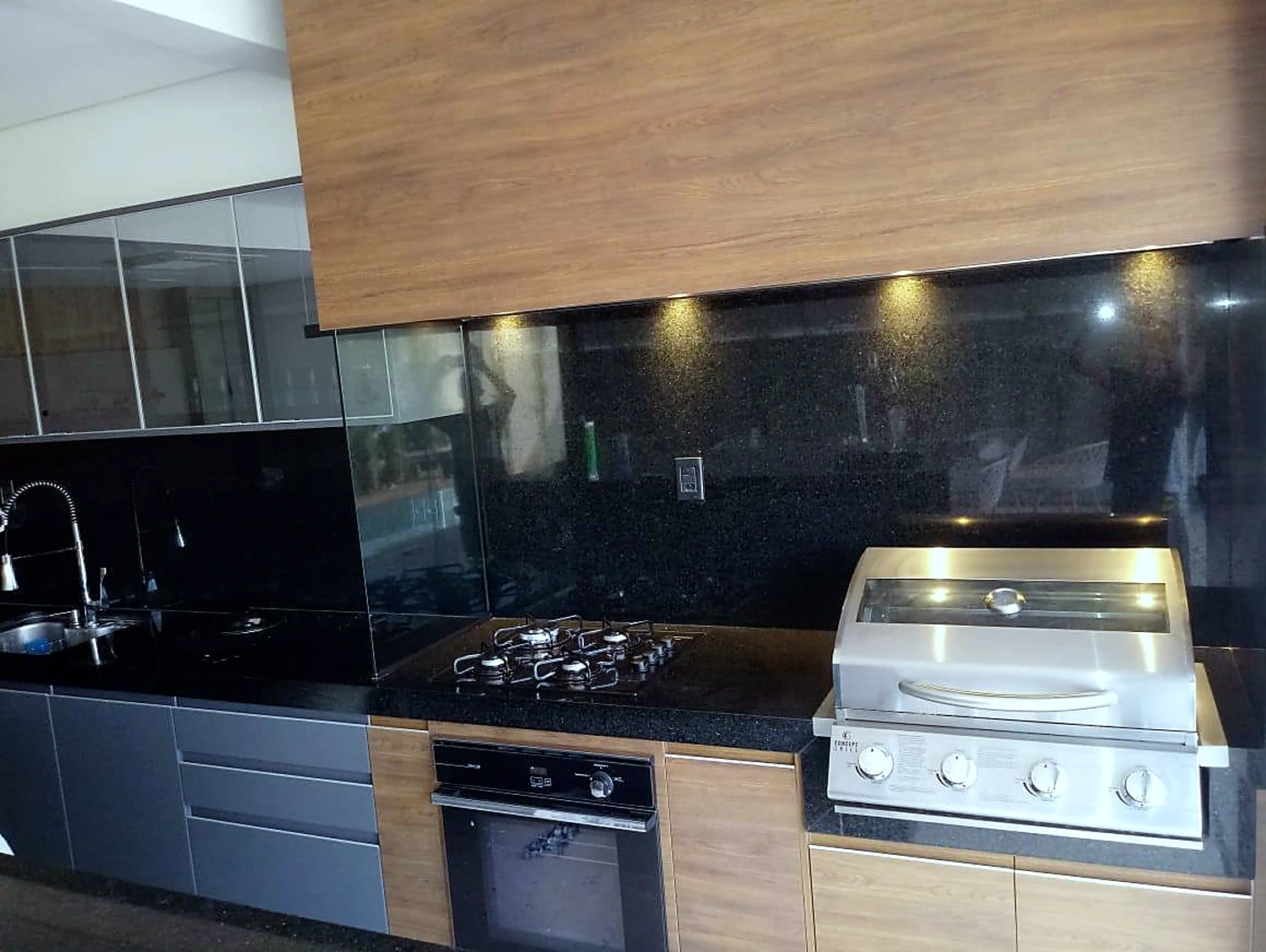 Área gourmet com churrasqueira em inox, cooktop embutido em granito preto, armários em madeira e iluminação sob o painel