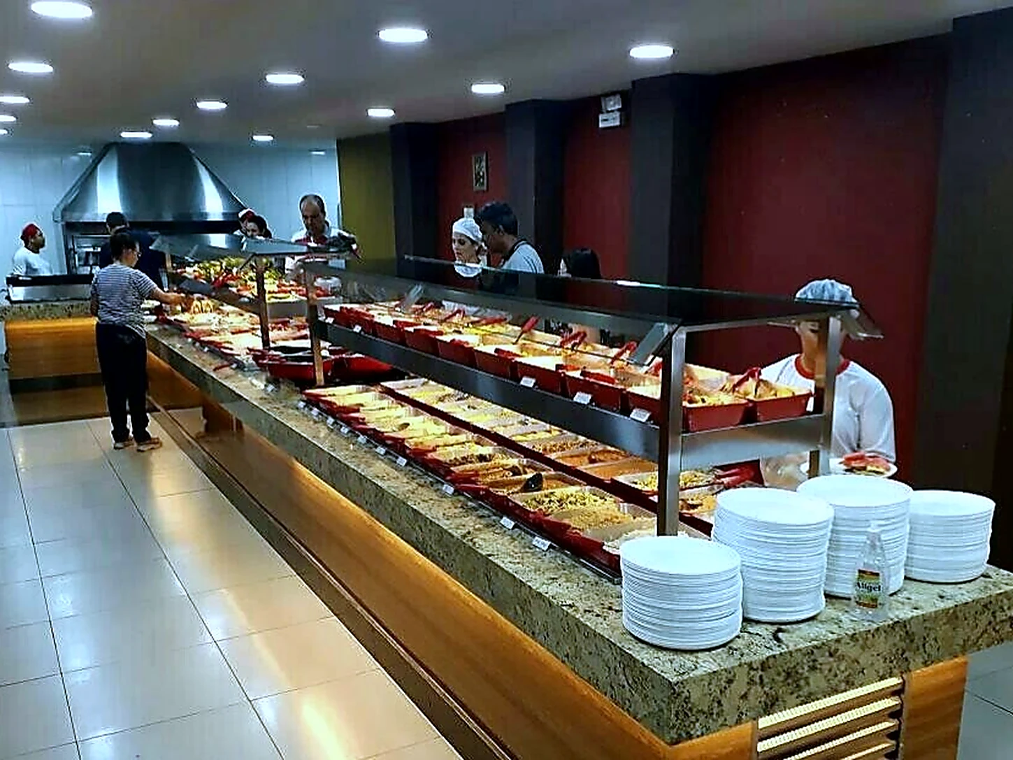 Linha de buffet de restaurante em plena operação, com cubas de alimentos, iluminação sobre a pista e equipe uniformizada atendendo