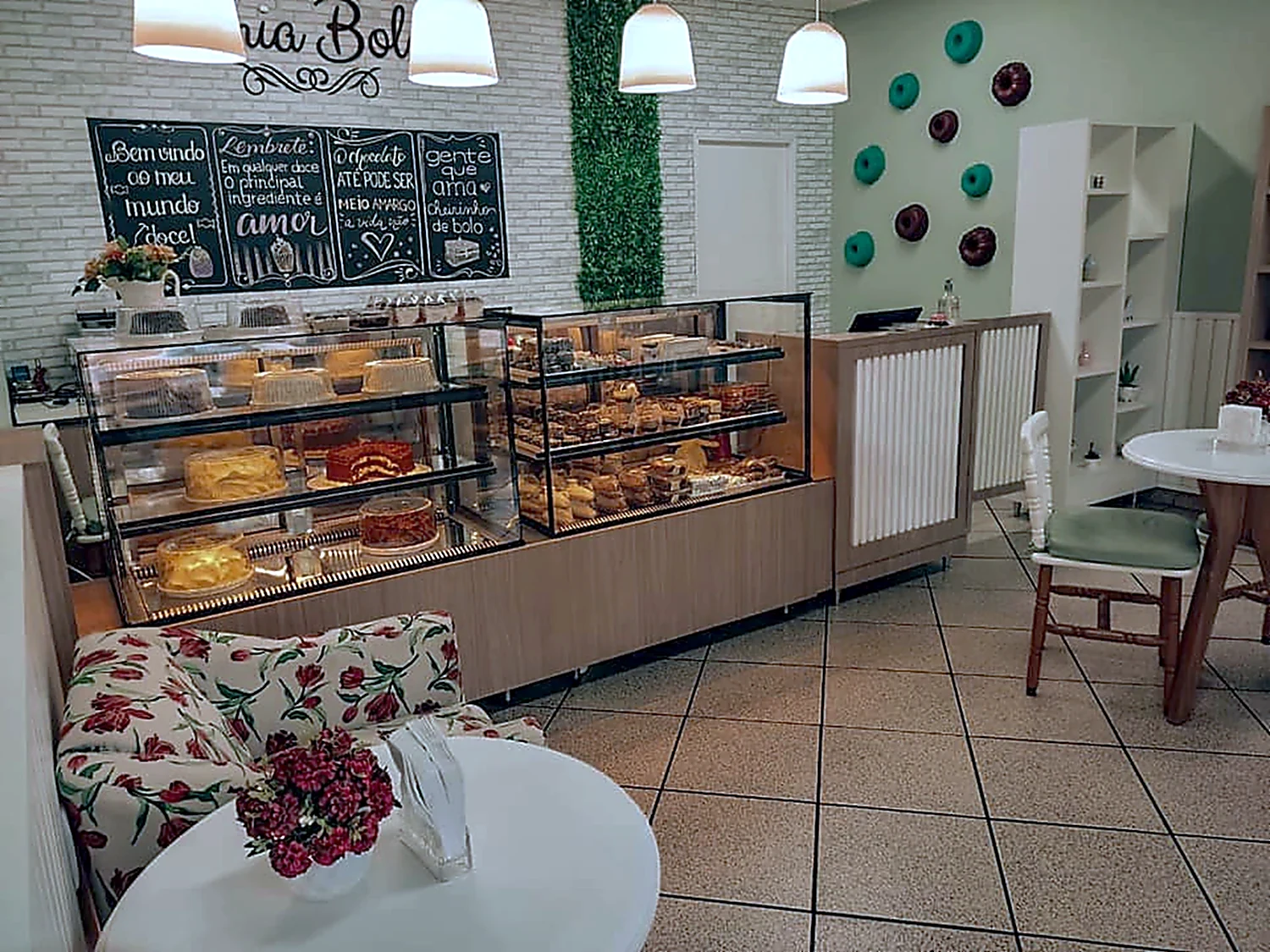 Balcão expositor refrigerado de uma cafeteria com pães e doces nas prateleiras, estrutura em madeira e salão montado ao redor